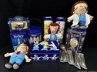 Group shot of six Tetley tins, three plush dolls, Lledo van in box, teaspoons in sleeves in front, tea strainer leaning on tin