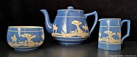 Front view of blue stoneware teapot, creamer and sugar bowl showing Tally Ho hunt scene relief
