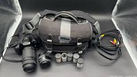 Lot overview showing Nikon bag, Minolta camera with Sigma lens, extra lens, film canisters, AV cable
