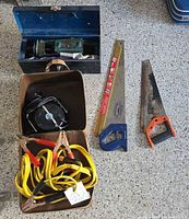 Lot overview showing jumper cables, cord reel in case, metal toolbox with bench vise, two hand saws