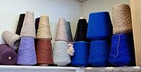 Shelf view of multiple yarn cones in assorted colours, left side includes purple cone lying horizontally and upright cones in greys, tans, blues and black