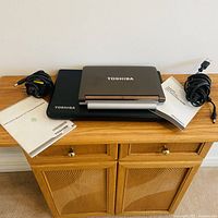 Two Toshiba laptops stacked with power adapters and manuals on cabinet
