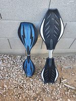 Two Razor RipStik caster boards leaning against concrete block wall, blue/black DLX Mini at left, black/white full-size at right