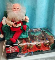 Front view of animatronic Santa seated on chair beside boxed Marching Band set