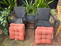 Full view of two wicker armchairs, square side table between, two orange cushions draped over fronts, garden backdrop