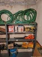 Shelf with green garden hoses, boxed utility pump, kettle, yellow sanders, leaf blower, jigsaw and water jug
