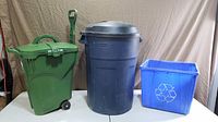 Front view of all three containers: green organics cart, black Rubbermaid Roughneck garbage can, blue recycling bin