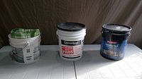 Three containers: white pail with green skim coat bag, white CertainTeed joint compound pail, black Black Knight foundation coating pail