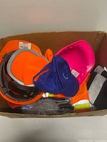 box containing orange Work+Play safety cap, goggles, blue cap, pink hard hat, hi-vis vest, black reflective garment, gloves packaged
