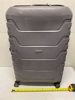 Front view of grey textured hard-shell suitcase on spinner wheels with tape measure indicating width (~19 in).