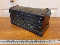 Front angled view of trunk showing wood construction, metal banding, latch, embossed floral panels, ruler for scale