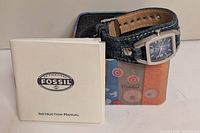 Watch with denim band resting on patterned Fossil tin alongside instruction manual