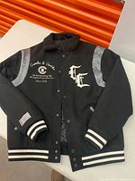 Front view of black varsity jacket showing logos, cuffs, waistband