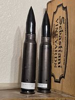 Two 30 mm dummy rounds standing upright beside a wooden box