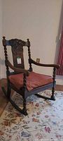 Front angled view of carved wood rocking chair showing barley-twist arms and legs, red cushion, dark brown finish