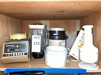Shelf view showing Osterizer blender base, blender jar, Moulinex chopper, Braun coffee grinder box, white bowls and attachments