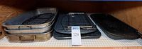 Stacked aluminum roasting pans with racks, two black broiler pans with racks, black grill pan