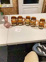 Five amber glass mugs and one pink bud vase on table