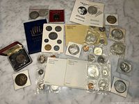 Overhead view of assorted foreign and commemorative coins, proof sets, presentation box, and flips displayed on marble surface