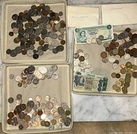 Three trays filled with assorted foreign coins and four banknotes placed on marble surface