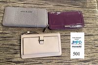 Three designer wallets laid out on fabric surface: grey Michael Kors studded zip-around, beige Kate Spade wristlet, purple Cole Haan patent wallet
