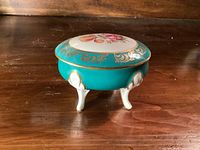 Closed trinket box showing turquoise body, gold trim, floral lid, tripod legs