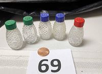 Front view of five pressed glass shakers with colored tops beside penny for scale