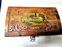 Top view showing curved lid, carved scrollwork, veneered landscape picture