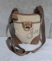 Front of Juicy Couture beige brown crossbody showing logo embroidery, leather trim, ring and tassel, adjustable strap