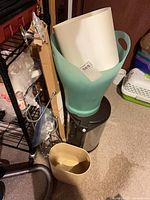 Group showing teal wastebasket inside white insert on top of stainless pedal bin, beige wastebasket in front, Moen kit bag on shelf, cat litter box to right