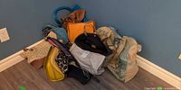 Pile of assorted handbags in corner showing orange tote, yellow bag, straw tote, blue handle bag, backpack and pouches