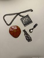 Four jewellery pieces laid out: square pendant on chain, enamel brooch, brutalist pin, tiny spool charm