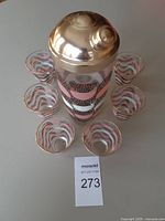 Shaker with lid surrounded by six glasses displaying matching pattern and gold rims