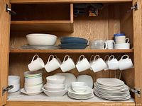 Cabinet view showing upper shelf: serving bowl, teal plates, glass measuring cup, assorted mugs