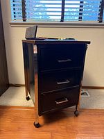 Cabinet side view showing black metal body, chrome frame, three drawers, casters