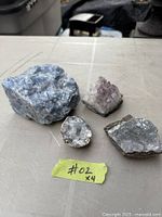 All four mineral specimens arranged on table: blue calcite, amethyst cluster, celestite geode half, grey stone thought to be kimberlite. Yellow tag #02 visible.