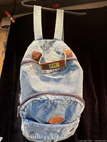 Front view of denim backpack showing main body, front zip pocket, leather patches and oval logo patch