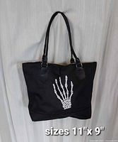 Front view of black nylon tote bag hanging, white skeleton hand graphic visible, size text 11" x 9"