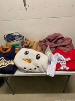 Table with assorted garments showing navy Chicago jacket, sherpa snowman hoodie, red hoodie, pink jacket, green costume, hangers