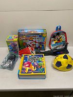 All items arranged on table including boxed marble run, boxed coin jar, backpack, ball, glove, cutlery, book, launcher parts