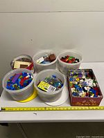 Six clear plastic tubs and one red floral metal tin filled with assorted building bricks shown on table with measuring tape