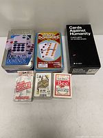 Group shot of all six items: two domino tins, Cards Against Humanity box, three card decks
