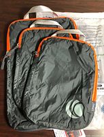 Three grey compression packing cubes stacked, showing sizes, orange zippers, front logo