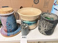 All four pieces grouped on countertop: stoneware jar with cork lid and saucer, beige planter, dark concrete planter with marbles, small terrazzo cylinder