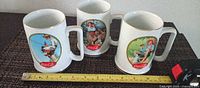 All three Coca-Cola mugs with tape measure showing height