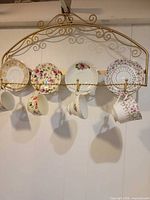 Full view of gold wall rack with six saucers slotted above and six teacups hanging below