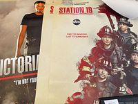 Two posters partially rolled showing Victoria Hughes character poster and ensemble cast poster side by side