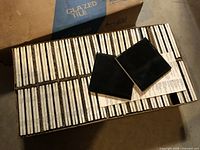 Open box showing rows of black glazed 4 x 4 in ceramic tiles with two tiles laid on top