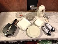 All lot items laid out: hand mixer with attachments, 2 TouCan openers, sandwich maker, plates, bowls, container