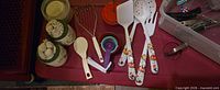 Utensils laid out on table: measuring cups, measuring spoons, wire whisk, rice paddle, three white utensils with fruit handles, ceramic canisters, plastic containers